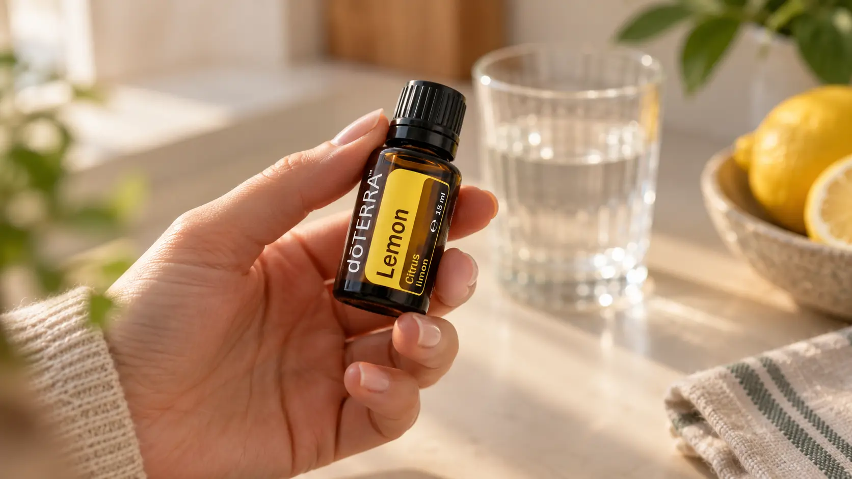 Dłoń trzymająca buteleczkę olejku cytrynowego doTERRA nad blatem ze szklanką wody i cytrynami